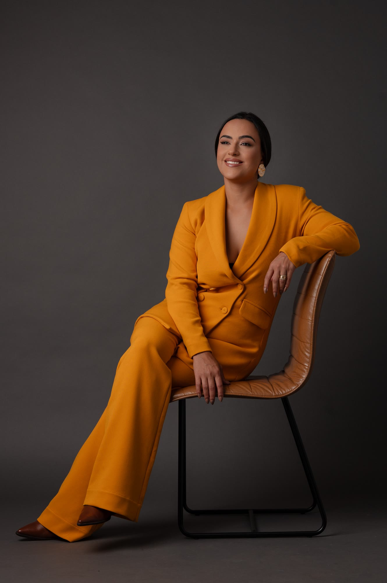 Camila Meneses sentada, de terno mostarda, em estúdio